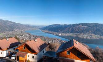 A panoramic view over a valley with a lake and surrounding mountains. In the foreground, several wooden houses can be seen.