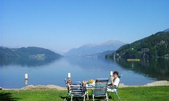 A family is sitting at a table by the shore of a tranquil lake. In the background, mountains and a clear blue sky can be seen.