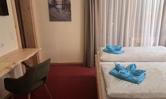 A cozy bedroom with two beds and blue towels. The room has a desk and large windows with curtains.