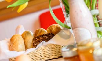 A woven basket with freshly baked bread and rolls. In the background, fresh flowers and a glass of yogurt can be seen.