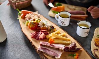A beautifully arranged plate with various types of sausages and cheeses, fresh vegetables, and a flower. In the background, cups with drinks can be seen.