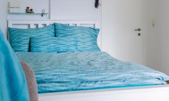 A cozy bedroom with a white bed and blue bedding. In the background, bright walls and a door are visible.