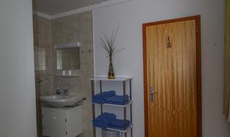 A simple bathroom with a sink, a mirror, and a wooden shelf. The wall is painted in light colors and there is a wooden chair.