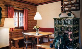 A cozy room with wooden walls and a table. In the foreground, there is a bouquet of flowers and a bag, while a traditional oven decorates the corner.