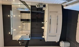 A modern bathroom with a stylish white sanitary installation and a large mirror. The walls are designed with dark tiles, and there are white towels as well as a trash can.