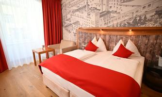 A cozy hotel room with a large bed and red pillows. The walls are decorated with a stylish wallpaper and large windows that let in plenty of daylight.