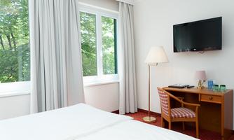 A bright hotel room with a double bed, a desk, and a television. Large windows let in plenty of daylight and offer a view of the greenery.