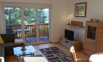 A bright living room with a cozy couch and a table.  
Large windows offer a view of a balcony and the nature outside.