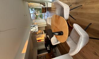 A modern dining area with a round wooden table and beige chairs. Large windows provide plenty of natural light and a view of the green surroundings.