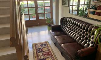 A cozy entrance area with a brown Chesterfield sofa and a beautiful rug. Large windows let in plenty of light, and plants create a pleasant atmosphere.