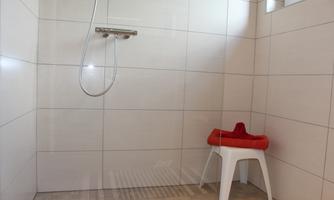A bright and modern shower with white tiles. In the corner stands a white plastic chair with a red towel.