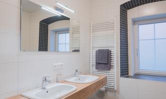 A modern bathroom with two sinks and a large mirror. There is a window with natural light and radiators next to the sinks.