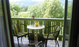 A cozy balcony with two chairs and a small table. The panoramic view shows green trees and a gentle hilly landscape in the background.