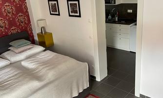 A cozy bedroom with a double bed and a beautiful carpet. In the background, a small kitchenette is visible.