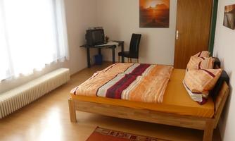 A simple bedroom with a large bed and colorful bedding. There is a desk, a chair, and a picture on the wall.
