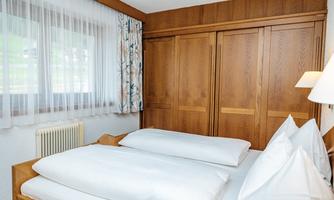A cozy bedroom with a bed, white pillows, and a wooden wardrobe. The room has large windows with curtains and is bright and cheerfully designed.