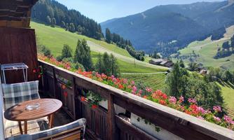 A balcony with a table and chairs, from which one has a picturesque view of green hills and mountains. Colorful flowers adorn the balcony railing.