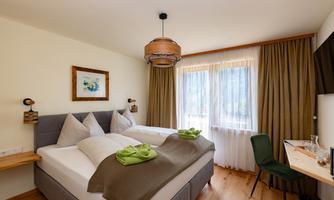 A cozy bedroom with two beds, a table, and a chair. Large windows let in plenty of daylight.