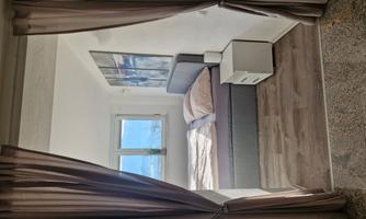 A cozy bedroom with a large bed and an open design towards the back. Natural light streams in through a window, creating an inviting atmosphere.