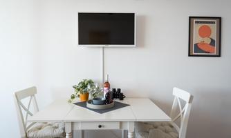 A bright room with a white table and two chairs. A television and a piece of art hang on the wall.