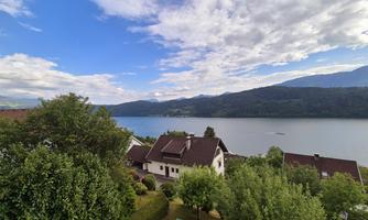 A picturesque landscape with a calm lake and gentle hills in the background. The surroundings are bordered by green trees and pretty houses.