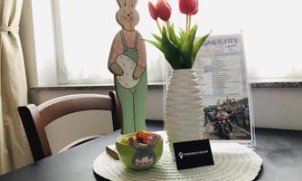 A decorative tabletop featuring an Easter bunny and a vase full of red tulips. Next to the vase is a bowl of sweets and an information card.