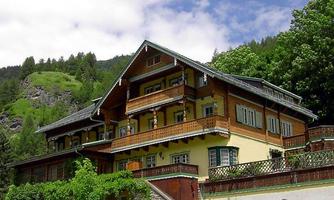 A traditional wooden house nestled in a green landscape. The facade features several balconies and surrounding trees.