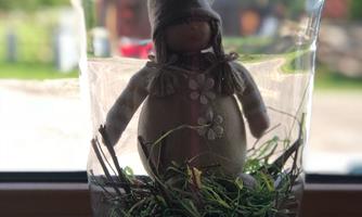 A decorative figure in a glass with grass and small flowers. The figure is wearing a gray hat and stands on a natural base.