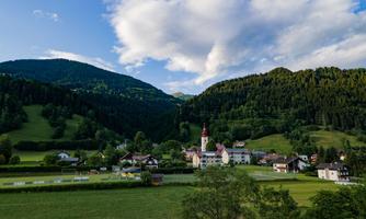A picturesque village view with a church surrounded by gentle hills. The landscape is green with trees and a clear sky.