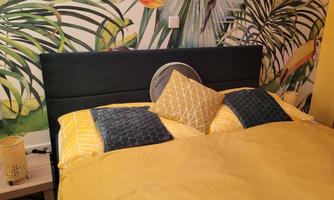 A cozy bed with yellow bed linen and decorative pillows. In the background, there is a vibrant wall with plant motifs.
