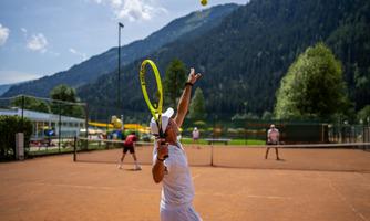 A tennis court in the midst of a picturesque landscape with mountains. A player is serving, while other players are active in the background.