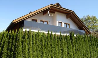 A modern house with a balcony and a sloped roof. The garden is surrounded by tall, green hedges.
