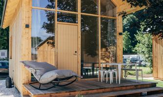 A modern wooden holiday home with large windows and a terrace. Outside, there are comfortable furniture and the surroundings are green and inviting.