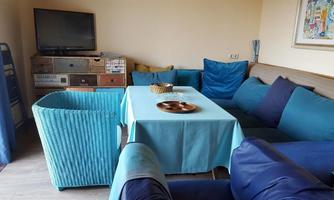 A comfortable living area with a blue couch and a table with a wooden top. In the background, there is a television and colorful cushions.