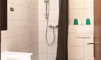 A small bathroom with a shower, a sink, and a toilet. The walls are tiled in light colors, and there is a black shower curtain.