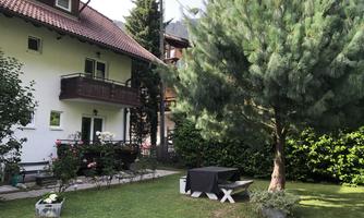 A well-maintained garden with a large tree and an outdoor table. The house in the background has a balcony and is surrounded by green plants.