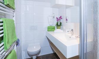 A modern bathroom with a white vanity unit and a shower. Green towels and a plant add a fresh touch to the space.