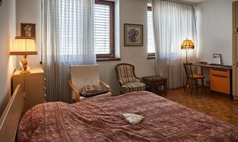A cozy bedroom with a large bed and a wooden chair.  
Large windows let in plenty of daylight, and a desk is in the corner.