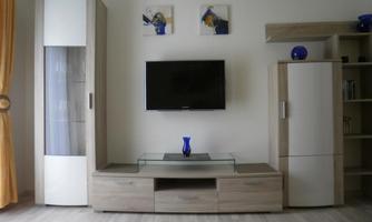 A modern living room with a TV, a glass table, and furniture in a light wood tone. Two pictures are hanging on the wall, and a blue vase is on the table.