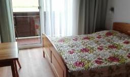 A simple room with a bed, a table, and a balcony. The room has large windows and a view of nature.