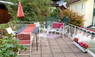 A cozy terrace with white furniture and a red sunshade. In the background, there are plants and a few flowers in boxes.