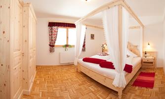 A bright bedroom with a wooden four-poster bed and red accents. The room has a warm wooden floor and friendly windows with curtains.