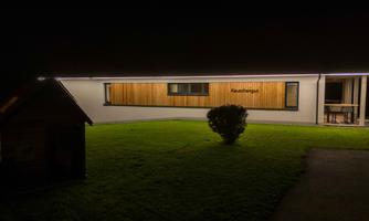 A modern building with a wooden facade and a illuminated veranda. The garden is well-maintained and the surroundings are dark.