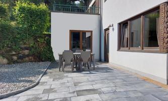 A cozy outdoor area with a table and chairs on a paved surface. In the background, a modern designed house and lush greenery can be seen.