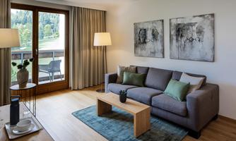A modern living room with a gray sofa and two cushions. Large windows provide plenty of sunlight and offer a view of the green surroundings.