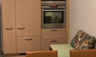 A modern kitchen with light wood cabinets and a built-in oven. In the foreground are two chairs and a table with a yellow tablecloth.