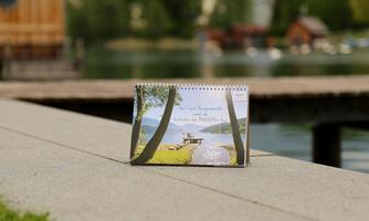 A calendar lies on a gray stone next to a calm lake. In the background, trees and a wooden terrace can be seen.