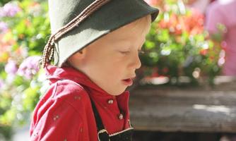 A boy in traditional clothing is working with wood. In the background, blooming plants and other people can be seen.