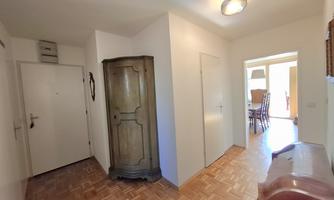 A bright entrance area with a wooden floor and a coat rack. To the right, there is an open room with a table and chairs.