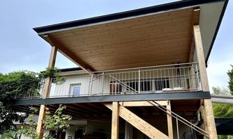 A modern house with a large covered wooden balcony. The construction is particularly distinguished by the striking supports and the surrounding greenery.